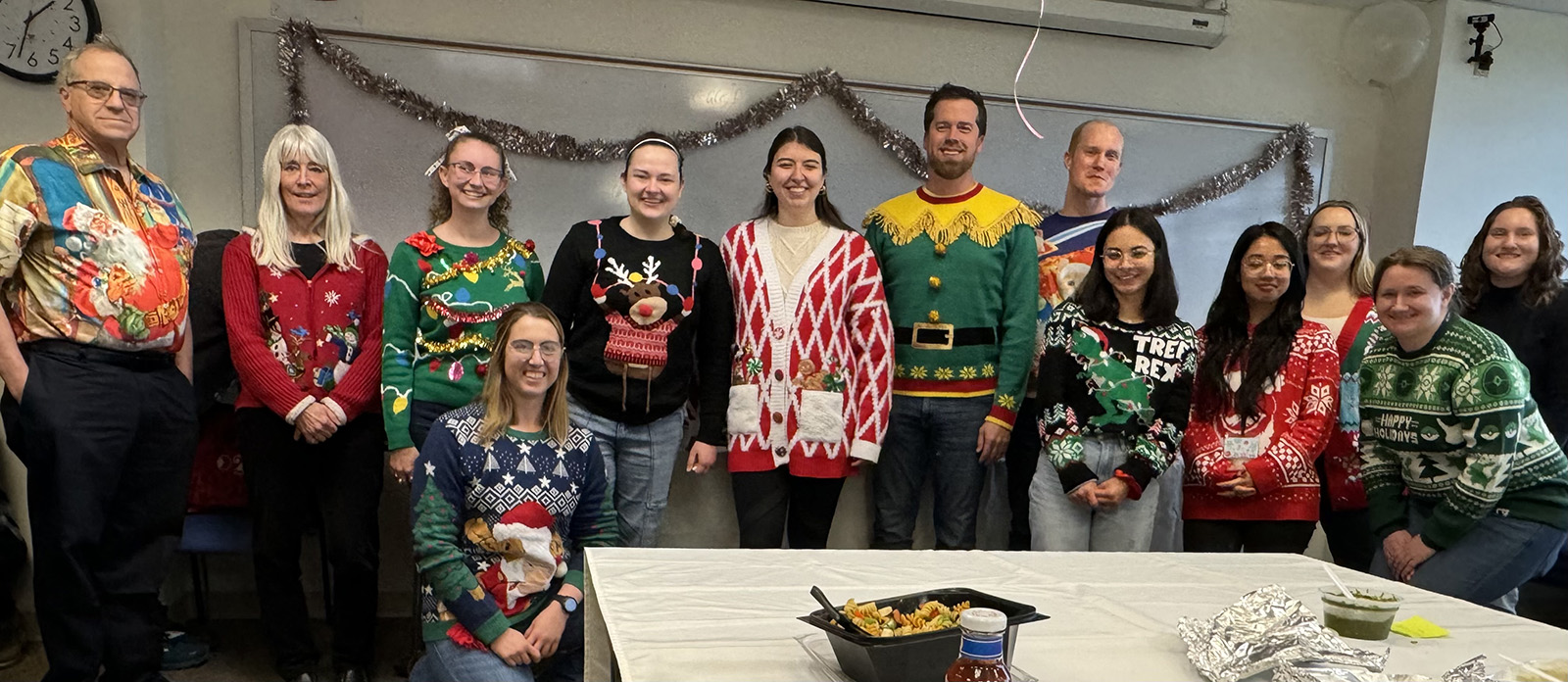 Thirteen people standing and one sitting in front, all wearing 'ugly' Christmas sweaters