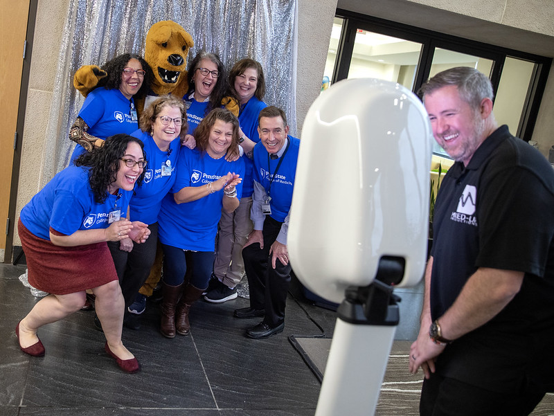 A group of seven people with the Nittany Lion mascot in the back poses for a photo booth picture with the photo machine and operator in the foreground. 
