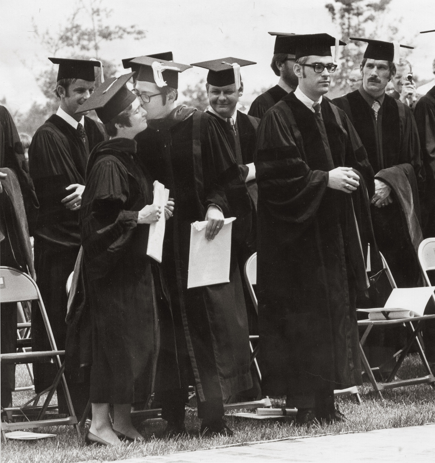About a half-dozen graduates in commencement gowns and caps stand among folding chairs; one kisses another on the cheek. 