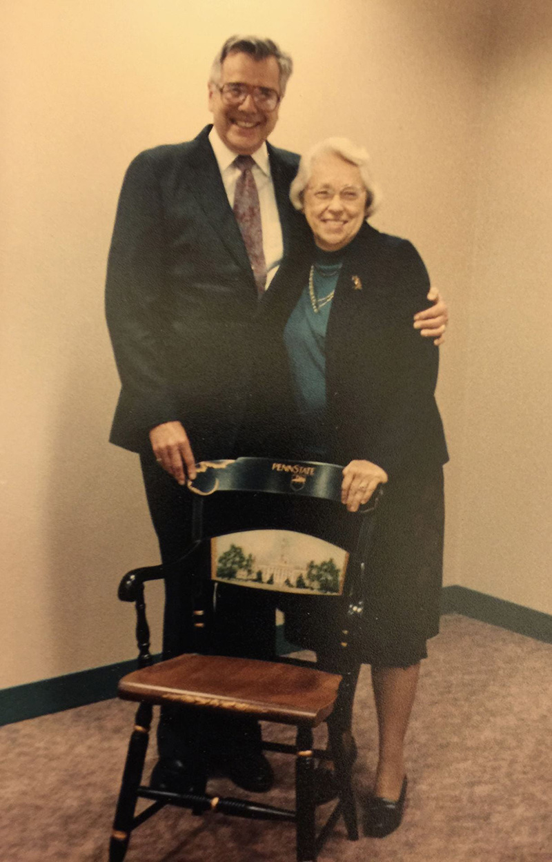 Lawrence F. Kienle, MD, and Jane Witmer Kienle, MD, stand with his arm around her and each holding onto the back of a wooden Penn State-themed chair.