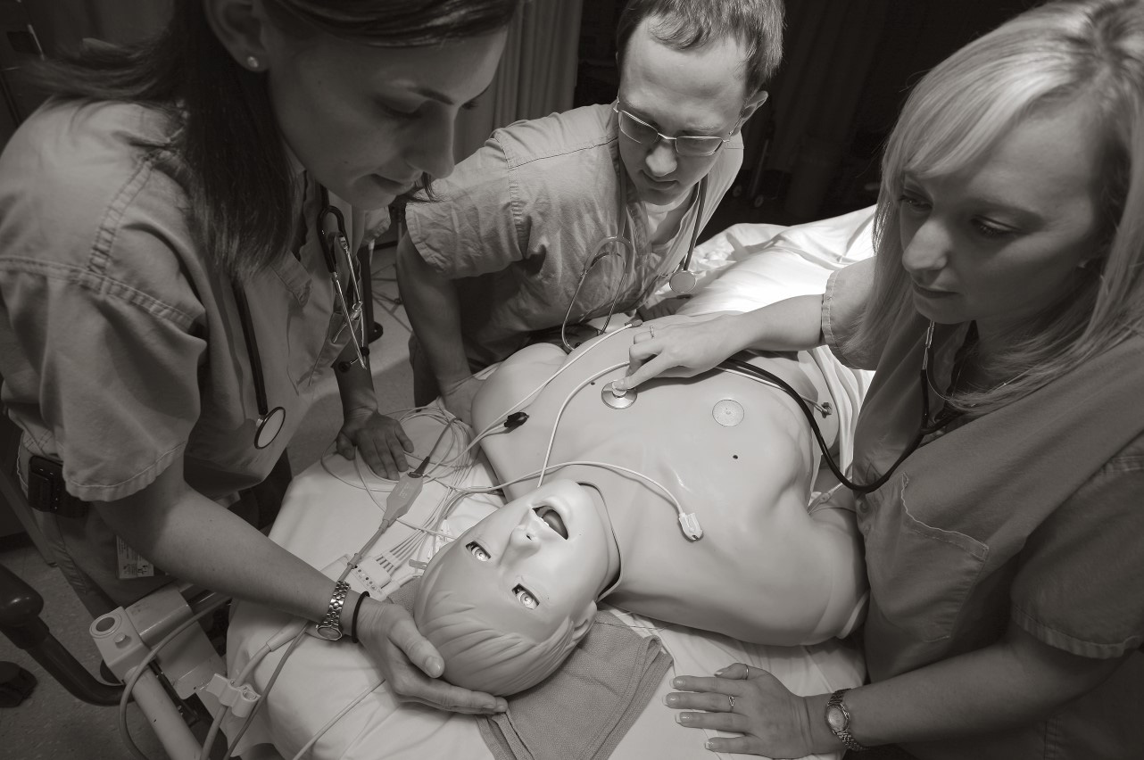  Three people stand around a mannequin; one holds a stethoscope to its chest while one holds the top of its head. 