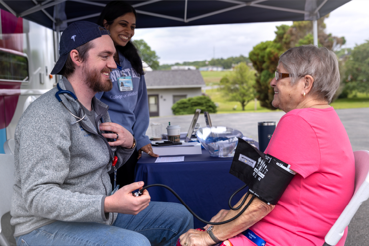 A medical student, seated, takes the blood pressure of a woman seated across from him while another student stands, smiling. They are under a rural mobile clinic tent.