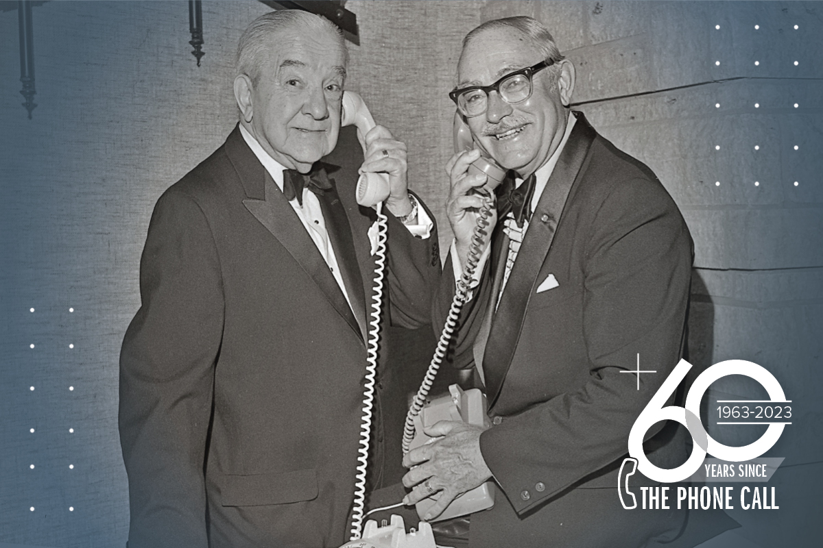 Sam Hinkle and Eric Walker each hold a corded phone to their ear in a recreation of the $50 million phone call in the 1960s; text in the bottom right corner says 60 years since the phone call, 1963-2023.