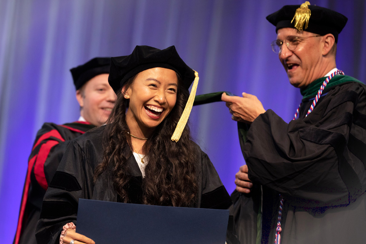 Lisa Ho smiles wide as she gets hooded by two faculty members in academic regalia during the 2023 Penn State College of Medicine commencement.