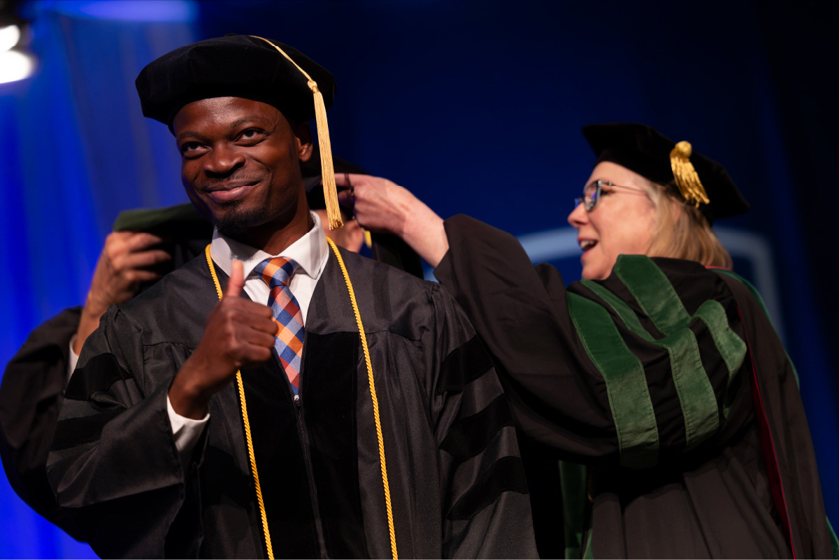 A graduate gives a thumbs up while a faculty member hoods him during the 2024 commencement at Penn State College of Medicine.