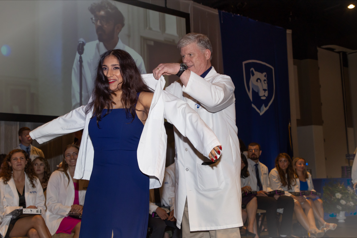 Dr. Michael Flanagan holds a white coat while an incoming medical student puts it on during the 2024 White Coat Ceremony.