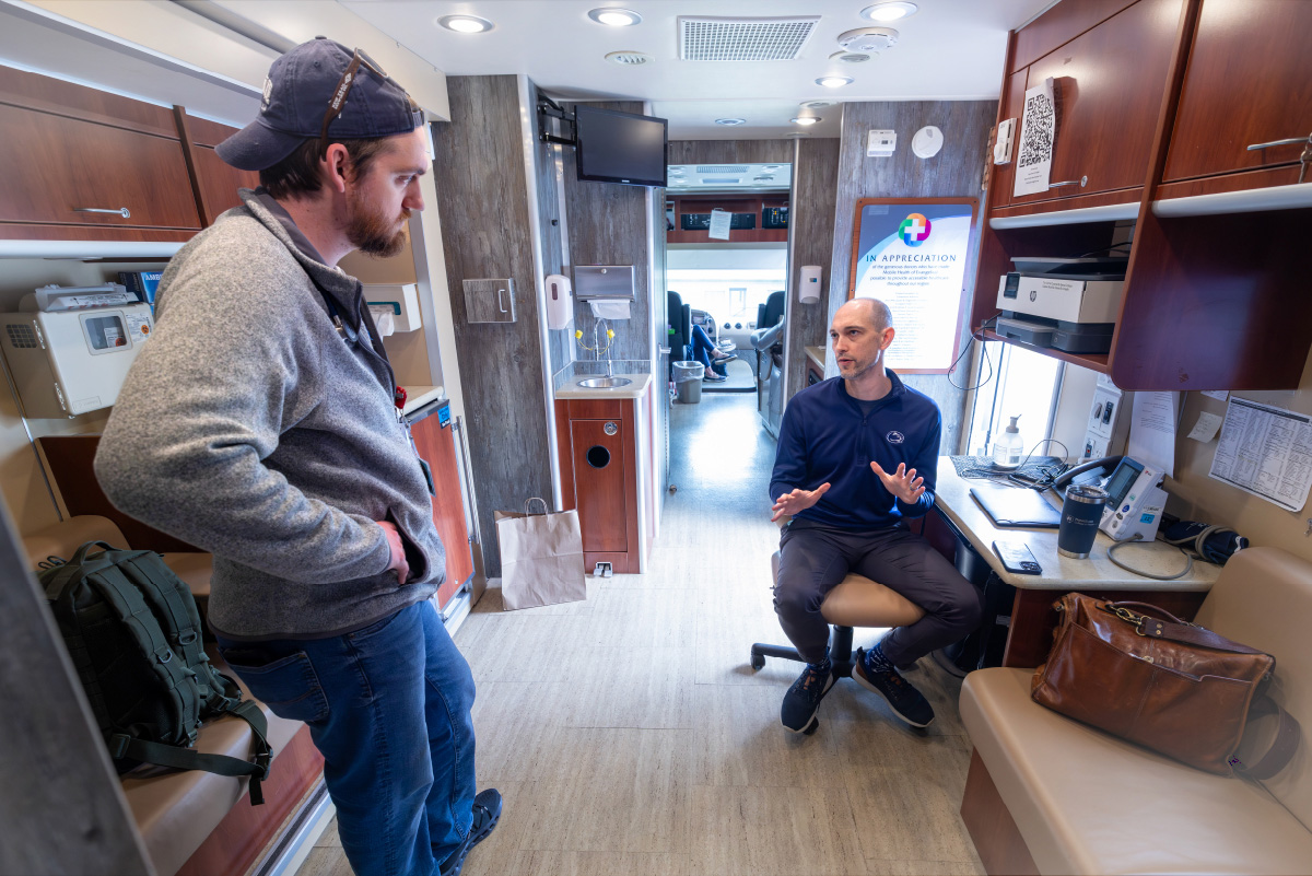 Dr. Michael McShane, seated at a desk, talks with medical student Derek Barnett, who is standing to McShane's right, inside the LION Mobile Clinic.