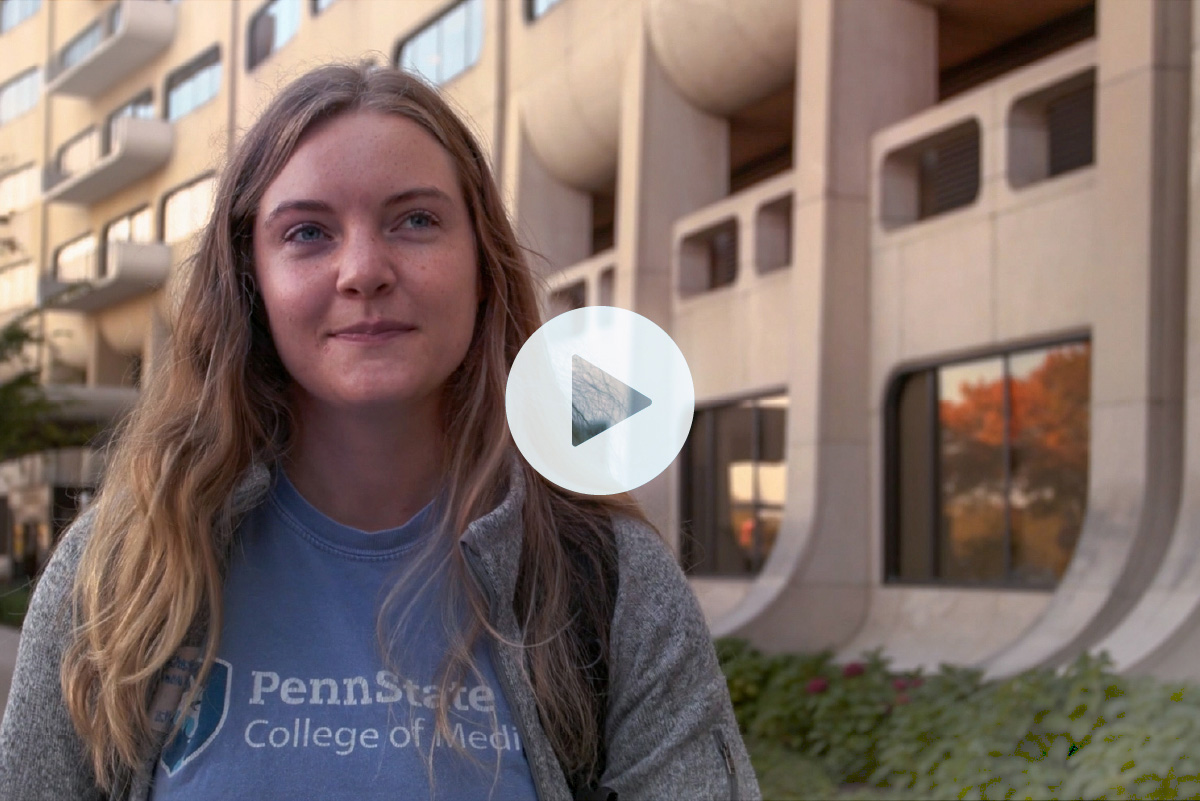 Lydia Smeltz wearing a Penn State College of Medicine shirt and standing in front of the college's crescent building, with a play button overlay.