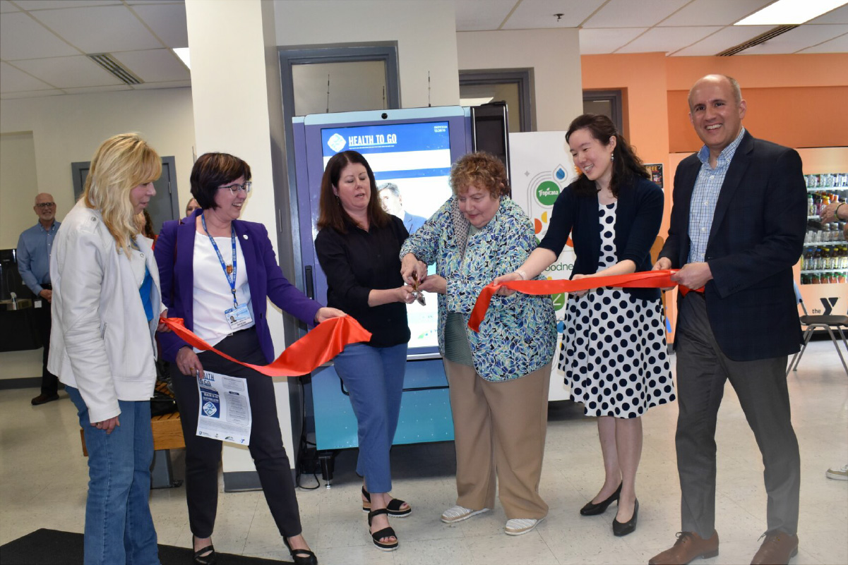 Four people, two per side, hold onto a ribbon while two people in the middle cut it during a ribbon-cutting ceremony in front of a Health To Go vending machine.