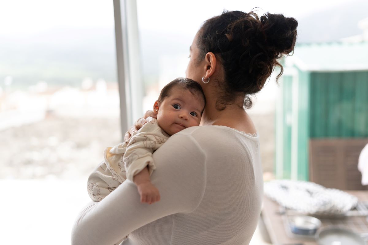 A woman holding a baby – the woman faces away looing out a window with the baby facing over her shoulder