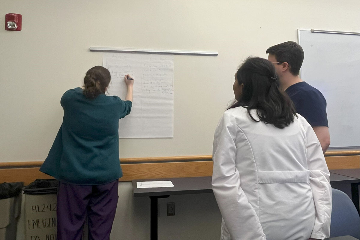 A person writes on a large piece of a paper hanging on a wall while two others watch