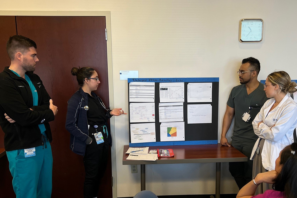 Four trainees stand by a table, two on either side, looking at a poster presentation